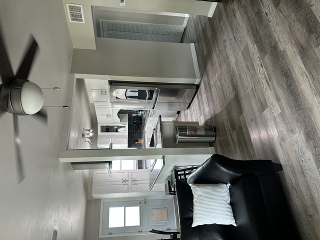 Living room and kitchen inside The Warriors Landing, with a leather sofa, wood-look floors, a ceiling fan, and a stainless-steel kitchen in the background